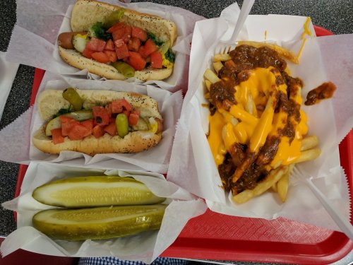 A red tray with two hot dogs topped with tomatoes and pickles, and a side of cheese-covered chili fries with pickles.