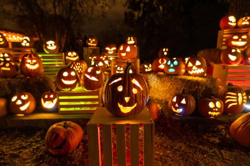 A festive display of carved pumpkins with glowing faces, surrounded by hay bales and a dark, spooky atmosphere.