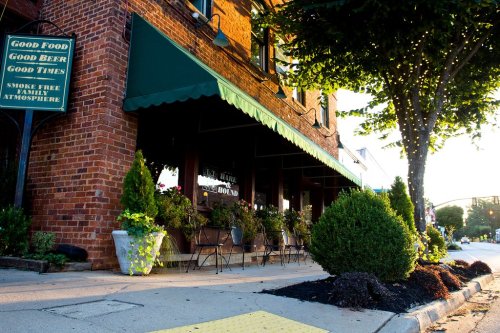 A brick restaurant with green awnings, outdoor seating, and potted plants, set on a sunny street.