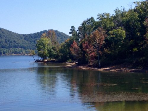 A serene lake surrounded by trees with autumn foliage, reflecting the clear blue sky and distant mountains.