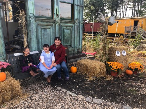 A woman and two girls sit outside a colorful building, surrounded by pumpkins, flowers, and hay bales in a festive setting.