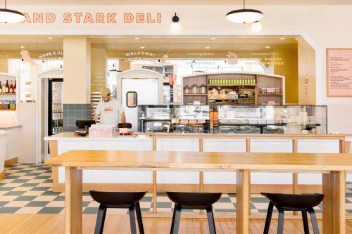 A bright deli interior with a counter, display case, and wooden tables and chairs. A staff member is behind the counter.