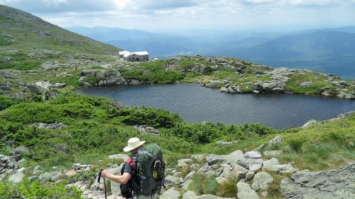 A hiker with a backpack descends a rocky path near a serene mountain lake, surrounded by lush greenery and hills.