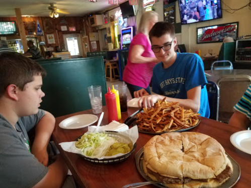Two boys sit at a table in a restaurant, one with a large burger and fries, the other looking on.