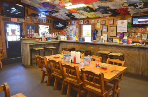 A rustic restaurant interior with wooden tables, bar stools, and a ceiling decorated with T-shirts and memorabilia.