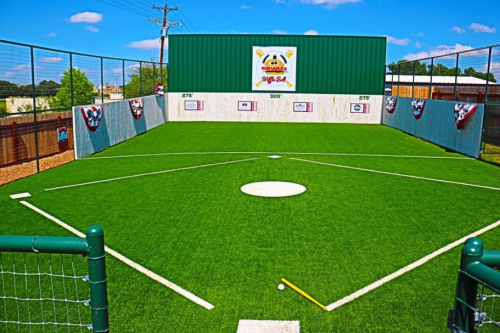 A small baseball field with artificial turf, featuring a home plate and a green wall in the background.