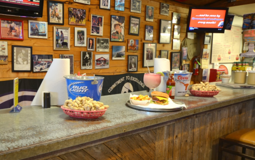 A rustic bar with a metal countertop, featuring drinks, snacks, and a burger, surrounded by framed photos on the walls.