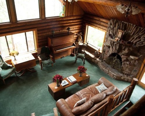 Cozy log cabin living room with a piano, stone fireplace, and comfortable seating, surrounded by large windows.