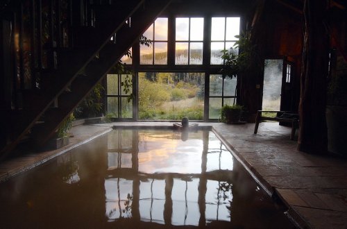 A serene indoor hot spring with a view of nature through large windows, reflecting a colorful sunset.