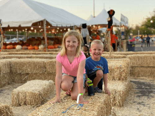 Two children play on hay bales at a pumpkin patch, smiling and enjoying the festive atmosphere.