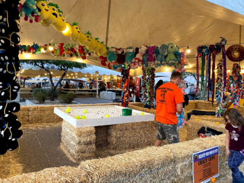 A festive fairground scene with a game area surrounded by hay bales, colorful decorations, and people enjoying the event.