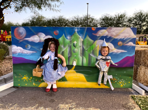 Two children pose in front of a colorful backdrop resembling the Emerald City, dressed as Dorothy and the Tin Man.