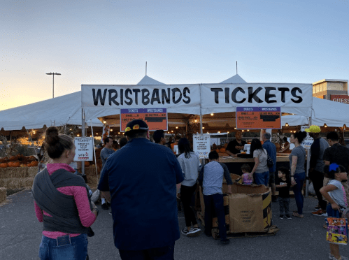 A busy ticket booth at an outdoor event, with people lining up for wristbands under a tent at sunset.