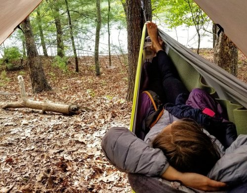 A person relaxes in a hammock between trees, surrounded by autumn leaves and a serene lake in the background.
