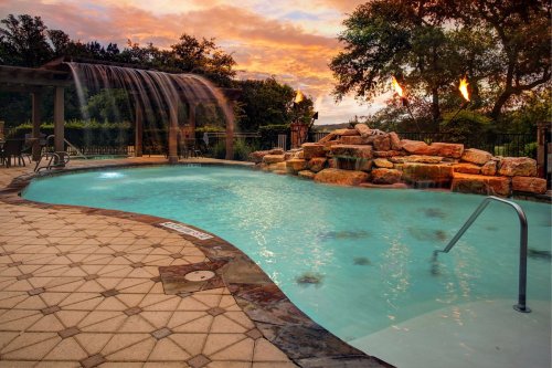 A serene pool with a waterfall, surrounded by rocks and a sunset sky, featuring lounge chairs nearby.