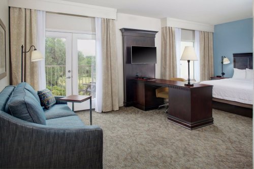 A hotel room featuring a bed, desk, sofa, and balcony doors with natural light and neutral decor.