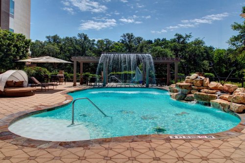 A serene pool area with a waterfall, lounge chairs, and lush greenery under a clear blue sky.