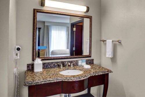 A hotel bathroom with a granite countertop, sink, mirror, and towel rack, featuring soft lighting and a window in the background.