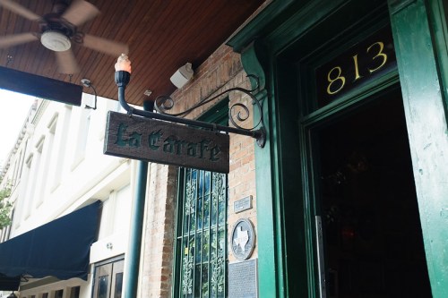 Sign for "La Carafe" hanging outside a brick building, with a green door and a decorative light fixture.