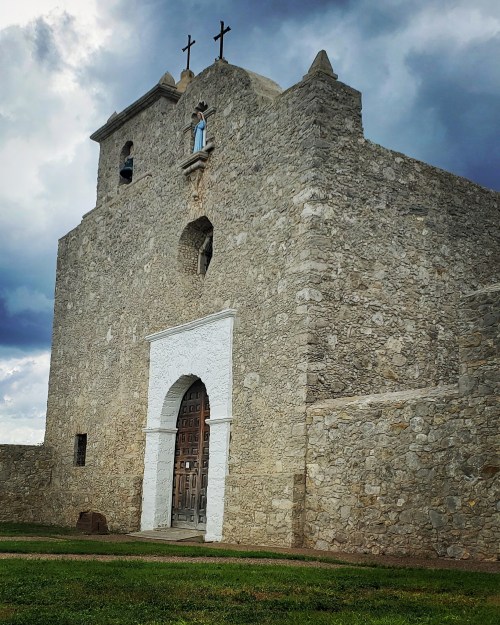 A stone church with a white archway and crosses on top, set against a cloudy sky. Green grass in the foreground.