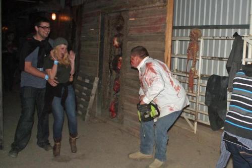 A man in a bloody shirt wields a chainsaw, while a woman appears startled and a man stands behind her in a dark setting.