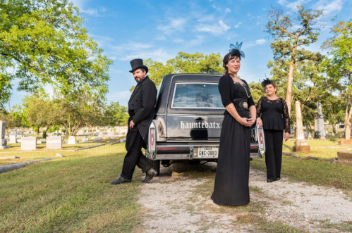 Three people in black attire pose near a hearse in a cemetery, surrounded by trees and gravestones under a blue sky.