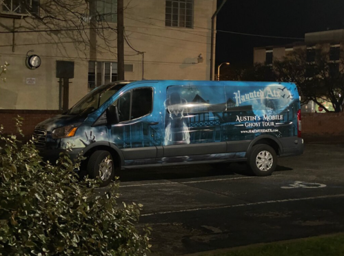 A blue van with a ghost tour advertisement parked in a lot at night, featuring a haunted theme and website details.