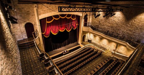 A grand theater interior with a red and gold curtain, empty seats, and stone walls, illuminated by soft lighting.