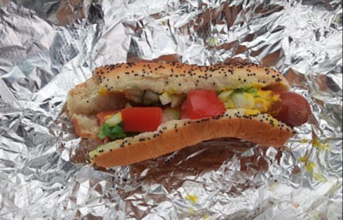 A hot dog in a poppy seed bun, topped with mustard, onions, tomatoes, and lettuce, wrapped in foil.