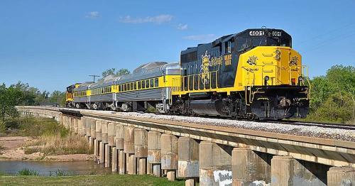 A yellow and black train crosses a bridge on a sunny day, surrounded by greenery and blue skies.