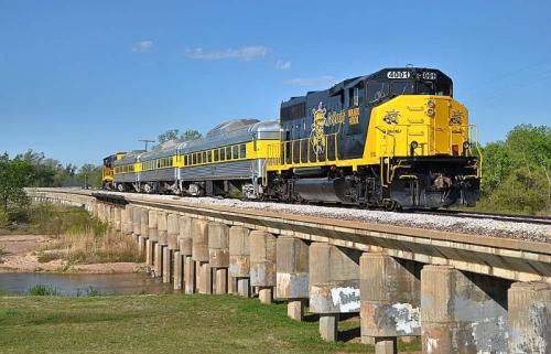 A yellow and black train crosses a bridge over a river, surrounded by greenery and a clear blue sky.