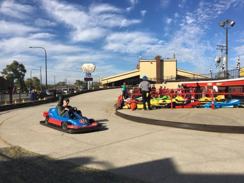 A go-kart on a track with a driver, while a referee watches nearby and colorful karts are parked in the background.