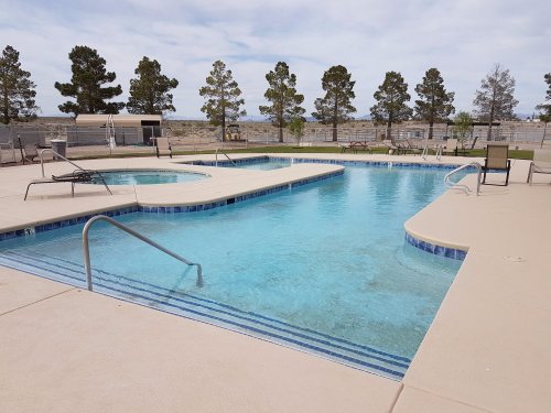 A clear blue pool with a hot tub, surrounded by lounge chairs and trees under a cloudy sky.