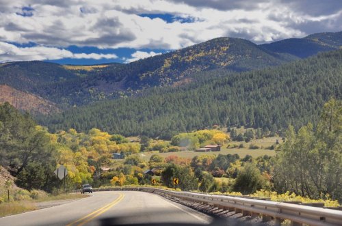 A scenic road winds through vibrant autumn foliage and rolling hills under a partly cloudy sky.