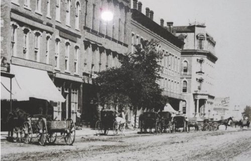 Historic black-and-white photo of a street lined with horse-drawn carriages and brick buildings.