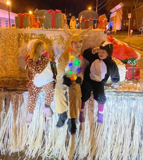 Three children in festive costumes sit on a decorated float, surrounded by holiday gifts and lights at night.