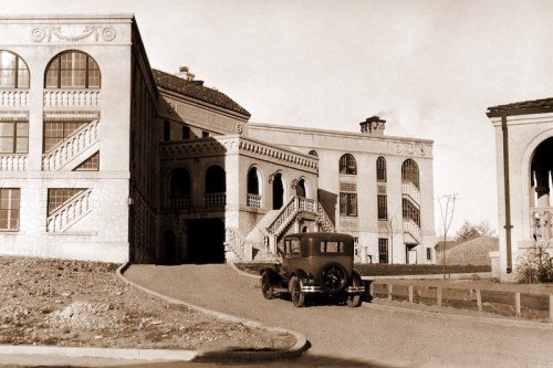 A vintage car parked near a grand, historic building with arched windows and staircases, set in a sepia tone.