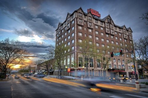 A historic hotel building with a peaked roof, surrounded by trees, under a dramatic sky at sunset. Traffic moves in front.