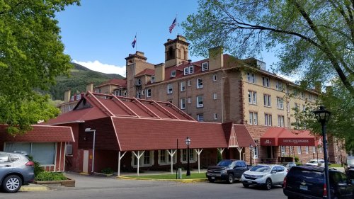 Historic hotel with red roofs and stone facade, surrounded by trees and parked cars, under a clear blue sky.