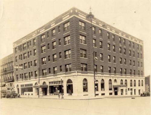 Historic brick building with multiple stories, featuring arched windows and a corner entrance, set in a vintage urban scene.
