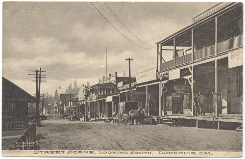 Black and white postcard of a street scene in Dunsmuir, California, featuring shops and horse-drawn carriages.