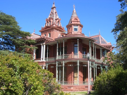 Victorian-style mansion with ornate towers and a wraparound porch, surrounded by greenery under a clear blue sky.
