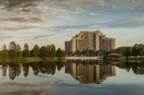 A modern hotel building reflected in a calm lake, surrounded by trees and a cloudy sky.