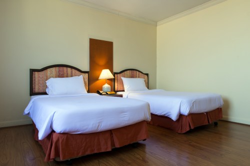 A bright hotel room featuring two twin beds with white linens and a bedside lamp on a nightstand.