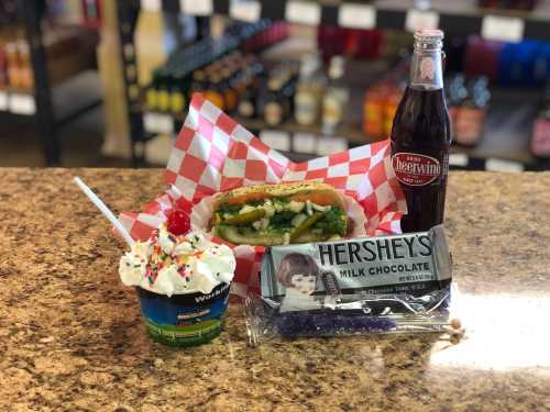 A hot dog with toppings, a chocolate bar, a cup of ice cream with a cherry, and a soda on a countertop.