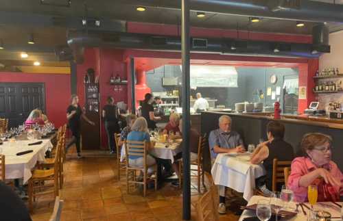 A busy restaurant interior with diners at tables and staff serving, featuring an open kitchen in the background.