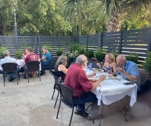 A group of people dining outdoors at a restaurant, surrounded by greenery and modern fencing.
