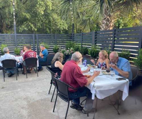 A group of people dining outdoors at a restaurant, surrounded by greenery and modern fencing.