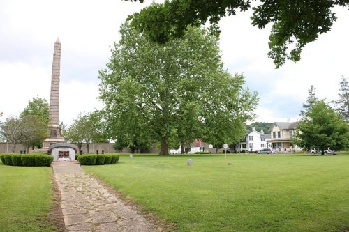 A grassy park featuring a tall obelisk, large trees, and historic buildings in the background under a cloudy sky.