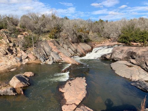 A serene landscape featuring a flowing river, rocky banks, and trees under a partly cloudy sky.